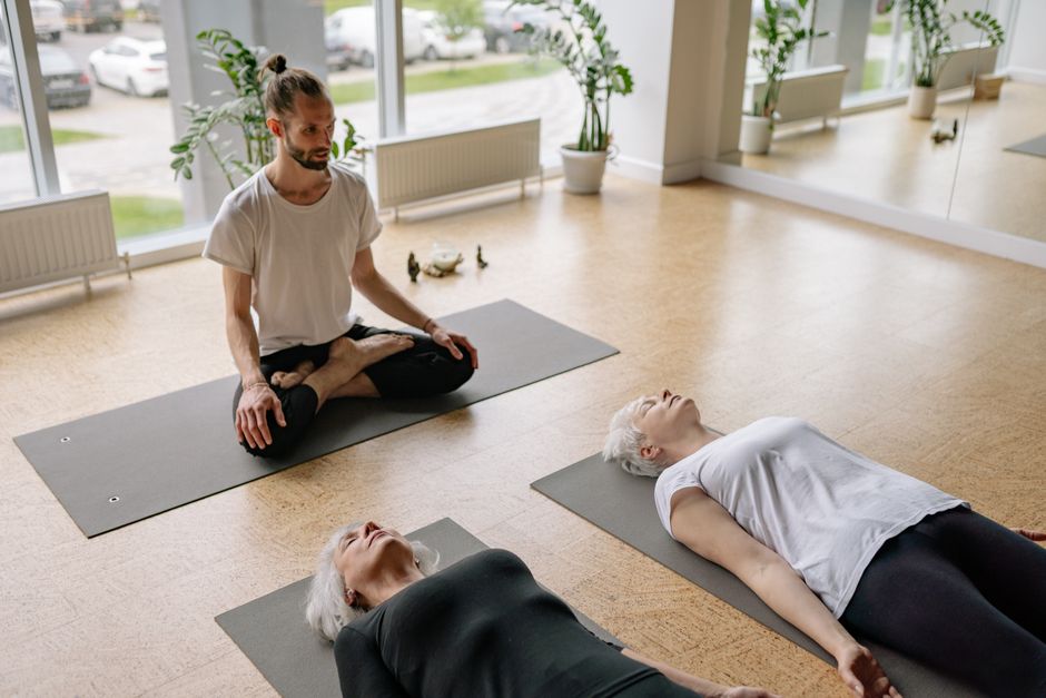 Pessoas participando de uma aula de ioga ou meditação em ambiente interno bem iluminado, com plantas decorativas e vista para o exterior.