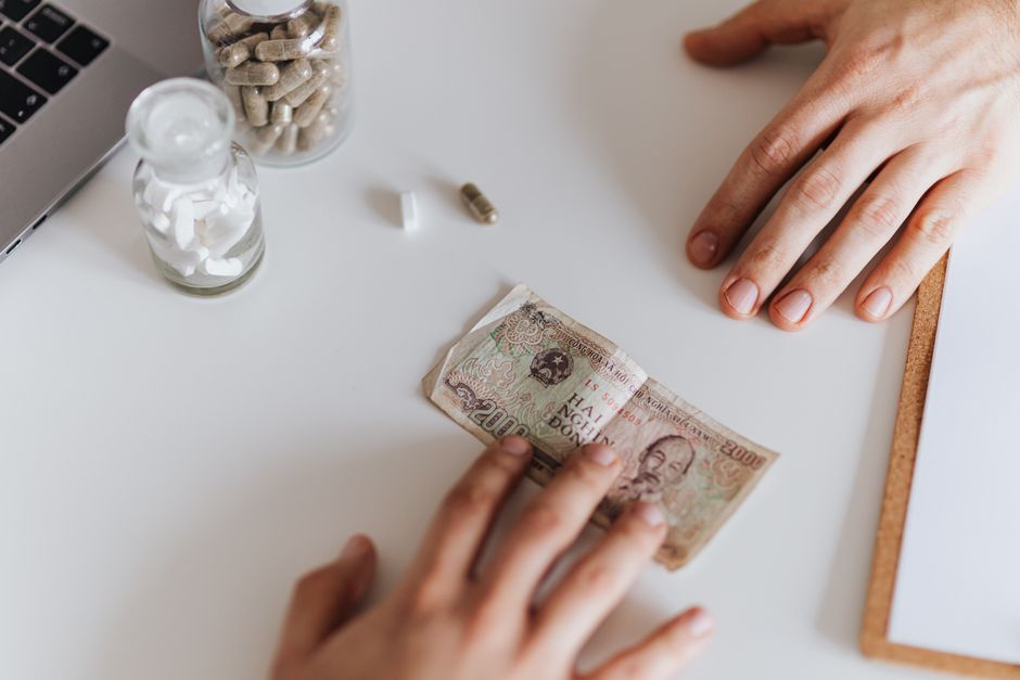 A imagem retrata uma situação de gerenciamento financeiro, com uma pessoa manuseando dinheiro em meio a medicamentos e um notebook, sugerindo temas de saúde financeira ou administração de recursos pessoais.