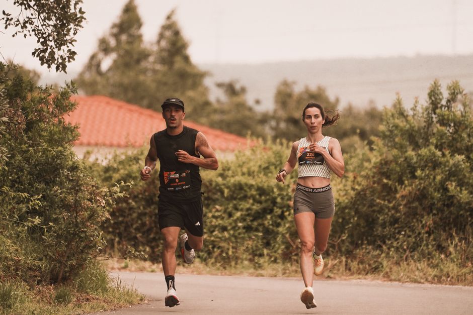 Duas pessoas correndo ao ar livre durante uma prova de corrida em um cenário Natural, com vegetação e céu nublado ao fundo.