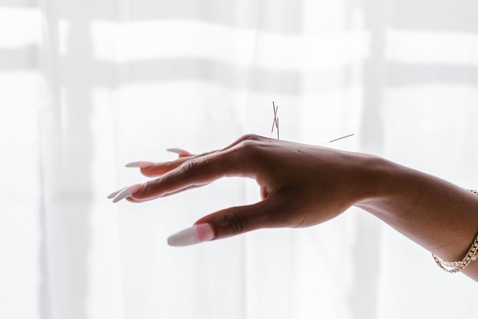 Mão feminina com unhas longas e bem cuidadas segurando agulhas de costura, simbolizando criatividade e artes manuais.