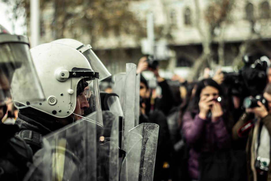 Policial em formação ou em operação, usando equipamento de proteção, com presença de fotógrafos ou jornalistas ao fundo, em um cenário urbano ao ar livre.