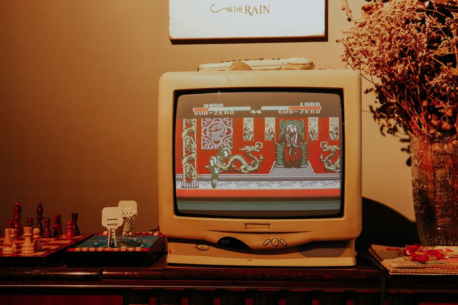 Imagem de um monitor antigo exibindo um jogo de arcade, com decoração aconchegante no ambiente ao redor, incluindo uma planta e um quadro na parede.