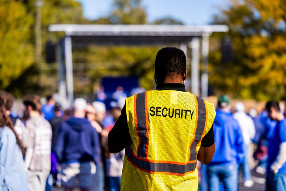 Imagem de um vigilante de segurança observando uma multidão em um evento ao ar livre durante o dia, com árvores ao fundo.
