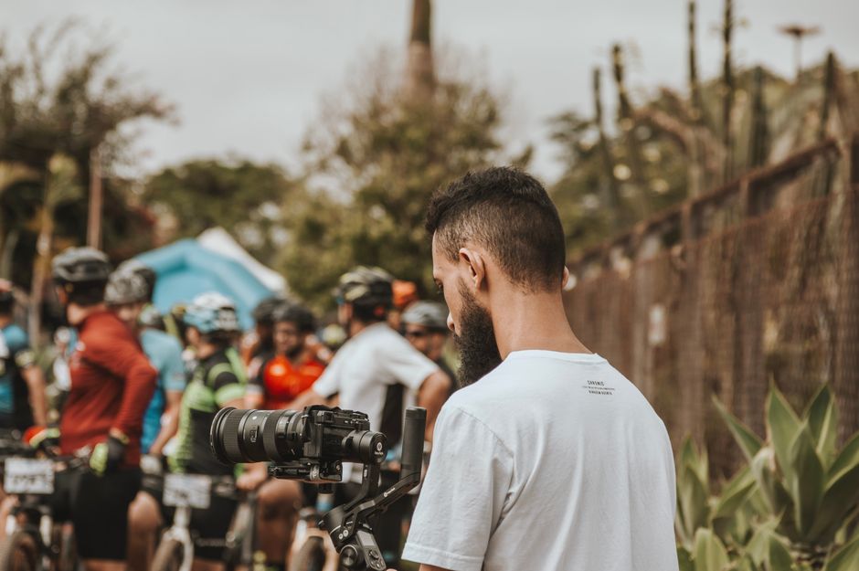 Homem ajustando uma câmera durante uma atividade de ciclismo ao ar livre com um grupo de ciclistas ao fundo.