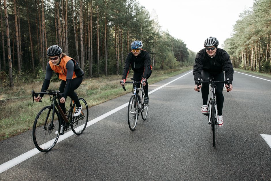 Três ciclistas praticando esporte ao ar livre em uma extensão de estrada cercada por árvores, refletindo saúde e lazer.