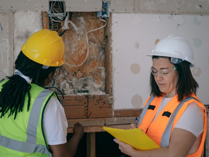 duas profissionais de construção civil analisando um projeto de obra em um ambiente de andaimes e paredes em construção