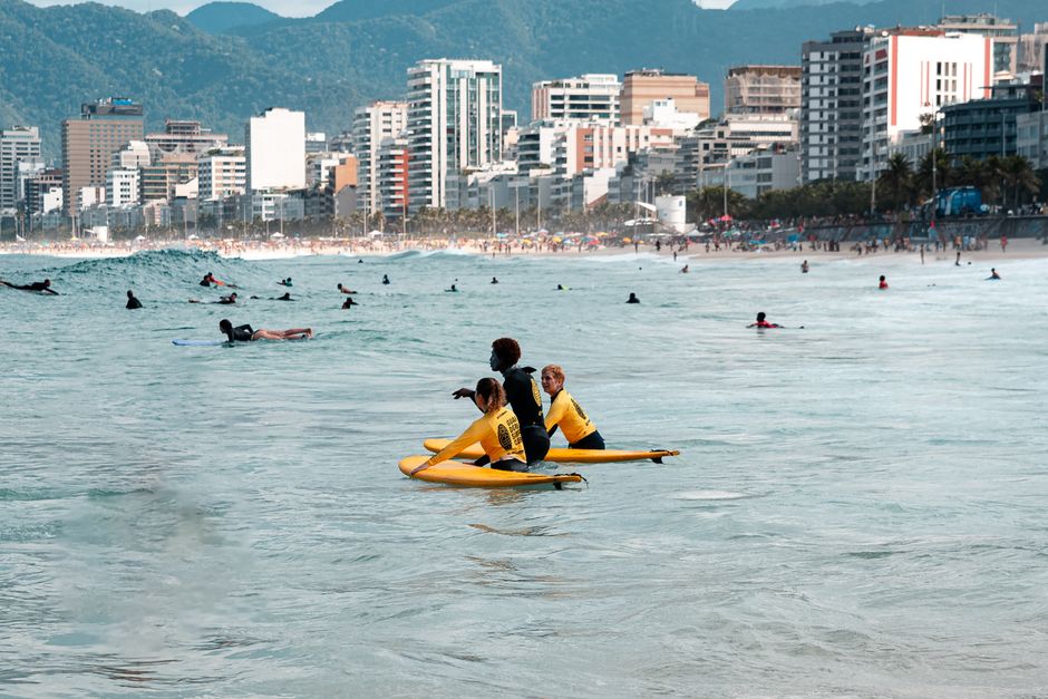Grupo de pessoas praticando surf na orla de uma cidade litorânea com arranha-céus ao fundo.