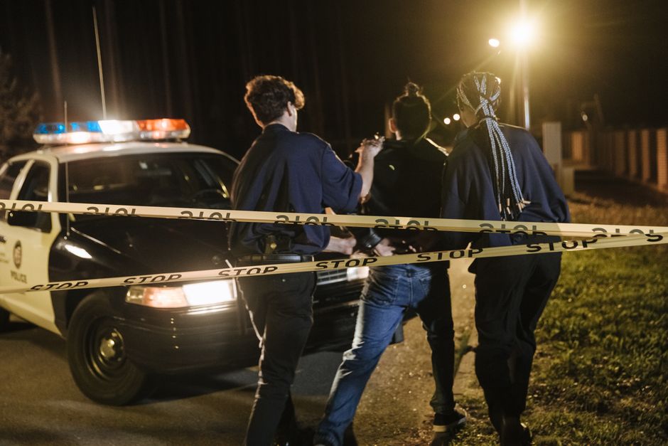 Três oficiais conversando na cena de um crime à noite, ao redor de uma viatura policial e com fita de isolamento na rua.