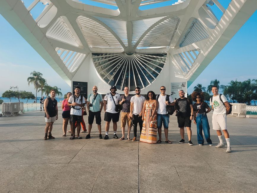 Grupo de pessoas posando em frente a uma estrutura arquitetônica moderna ao ar livre, com uma atmosfera descontraída e turística.
