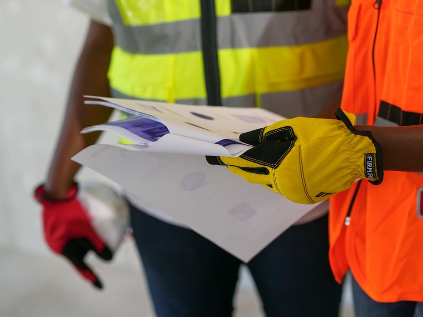 Pessoas usando coletes de segurança revisando documentos em um ambiente de construção ou obra, focando na segurança e planejamento do projeto.