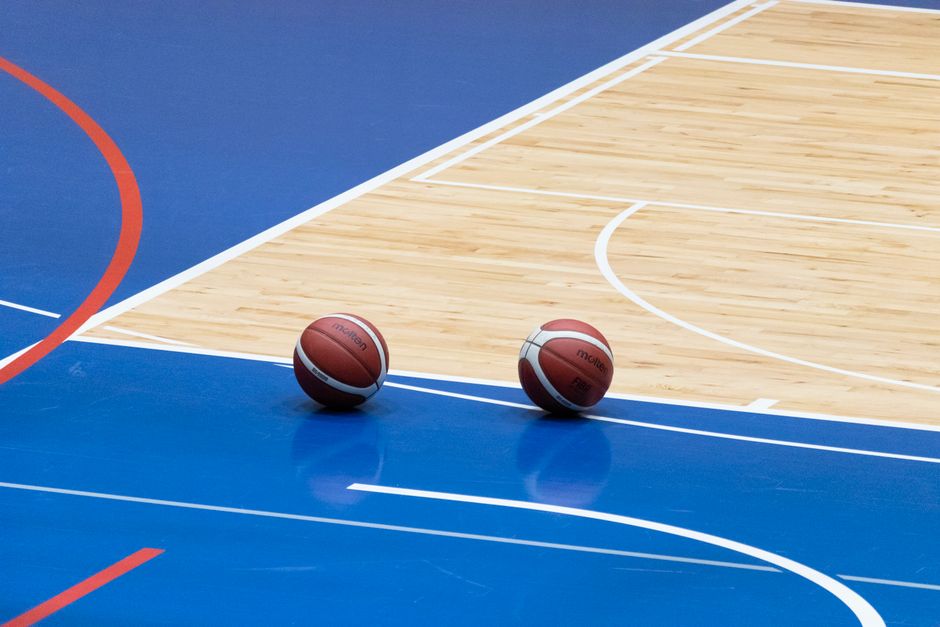 Imagem de uma quadra de basquete com duas bolas no piso, destacando o ambiente esportivo e o movimento do jogo.