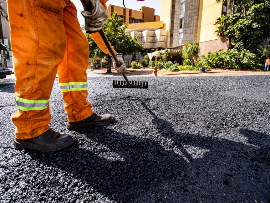 Imagem de um trabalhador em obras, com roupas de segurança, realizando o asfaltamento de uma rua em um ambiente urbano.
