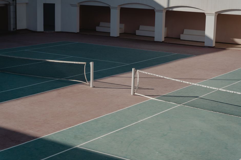 Imagem de uma quadra de tênis ao entardecer, com duas redes dividindo o espaço e um ambiente cercado por áreas cobertas e escadas ao fundo.