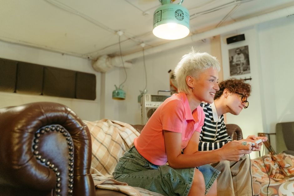 Duas pessoas, uma com cabelo curto e loiro e outra com cabelo cacheado e escuro, jogando videogame juntas em um ambiente descontraído, demonstrando diversão e socialização.