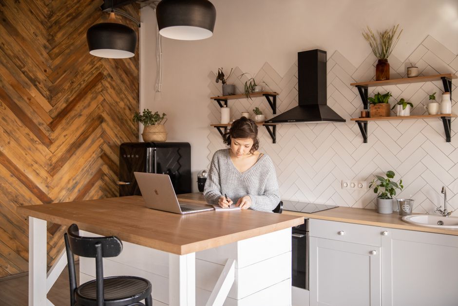 Mulher estudando ou trabalhando em um ambiente doméstico de cozinha moderninha, com tablet e laptop, em um cenário que combina funcionalidade e aconchego.