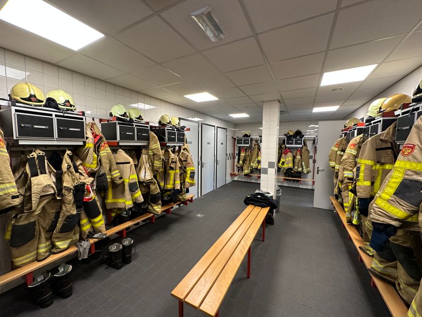 Sala de vestiário de bombeiros com uniformes, capacetes e equipamentos prontos para uso.