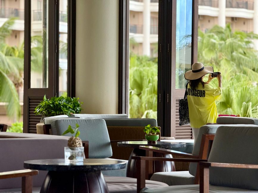 Pessoa de chápeu e roupas de viagem observando a paisagem tropical a partir de uma varanda de hotel.