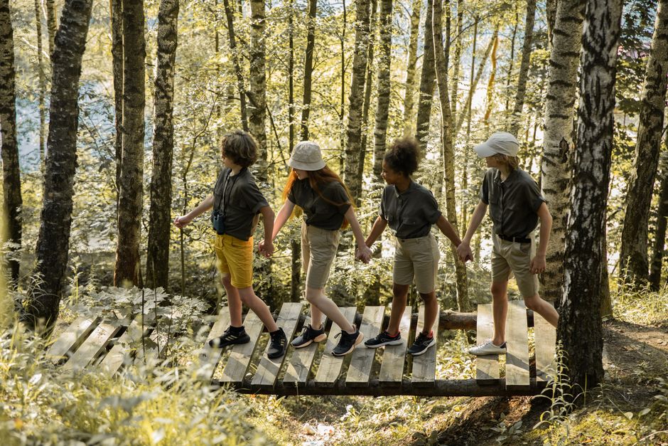 Grupo de crianças caminhando de mãos dadas em uma trilha de madeira na floresta, explorando a natureza.