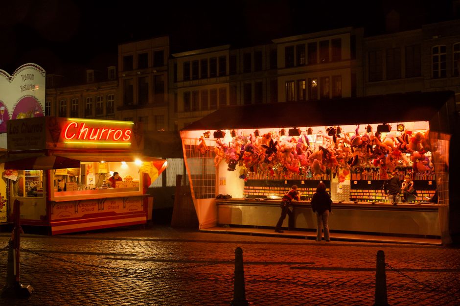Feira noturna ao ar livre com barracas de comida e jogos, iluminada por luzes quentes e destaque para os brinquedos de pelúcia no estande de diversão.