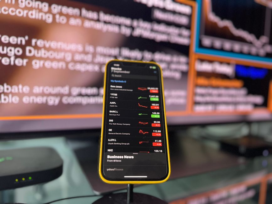 Imagem de um celular com tela exibindo indicadores de ações em um ambiente de notícias financeiras, com telas de gráficos de mercado ao fundo.