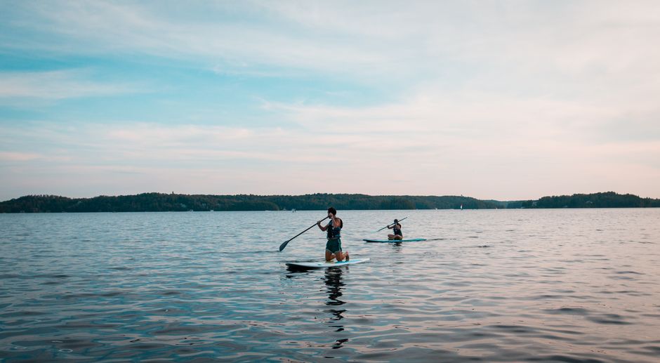Duas pessoas praticando stand-up paddle em um lago tranquilo sob um céu com algumas nuvens, em um cenário de lazer ao ar livre.