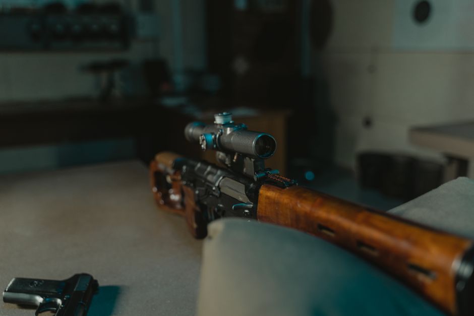 Imagem de um rifle com mira, exibido em um ambiente interno que parece ser uma sala de armazenamento ou armamento.
