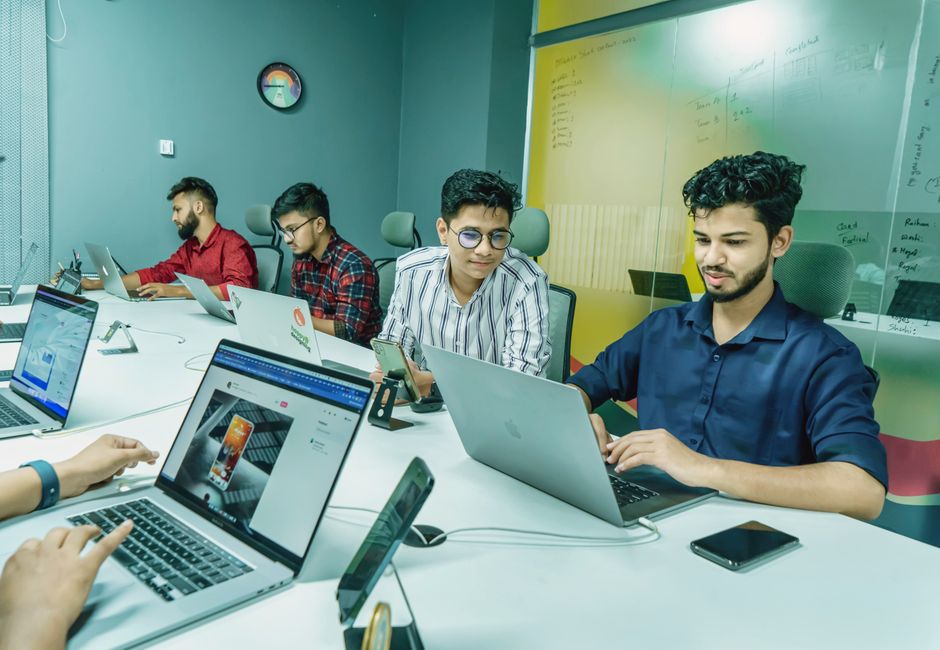 Grupo de jovens profissionais colaborando em uma reunião de trabalho em ambiente de escritório moderno, focados em tecnologia e planejamento.