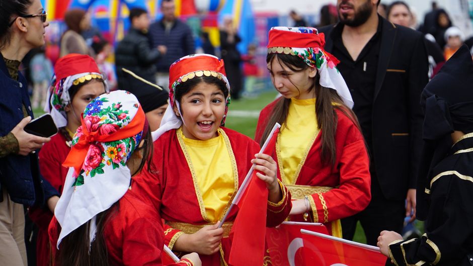 Grupo de crianças participando de uma festa cultural ao ar livre, vestidas com trajes tradicionais e segurando bandeiras.