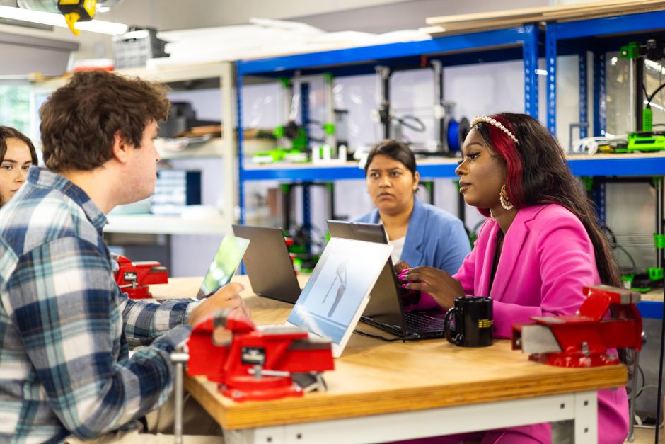 Grupo de jovens colegas conversando em um ambiente de inovação tecnológica com notebooks e impressoras 3D ao fundo.