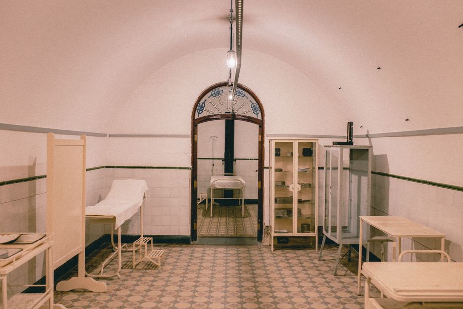 Sala de hospital desativada com móveis antigos e teto arqueado, evocando atmosfera de abandono ou repouso esquecido.