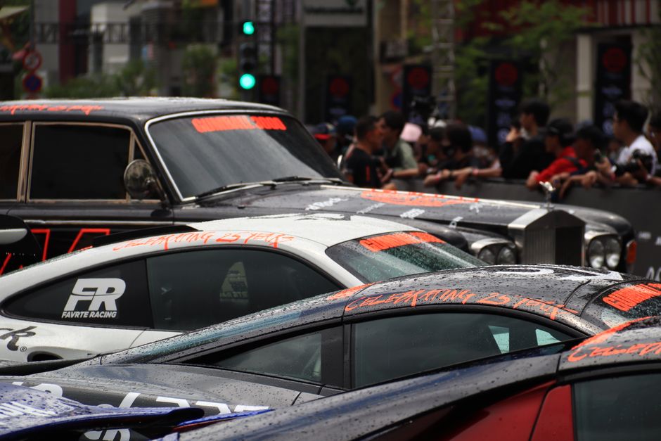 Carros de corrida parados em uma rua movimentada, com pessoas assistindo ao fundo, em evento de automobilismo urbano.