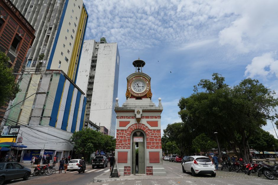 Imagem de um relógio de estação em uma praça urbana com prédios altos ao fundo e céu com nuvens dispersas.