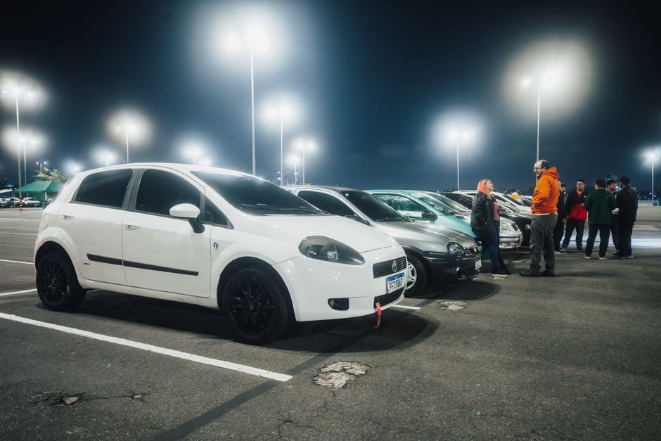 Grupo de pessoas conversando ao lado de carros em estacionamento, ao anoitecer, sob iluminação artificial.