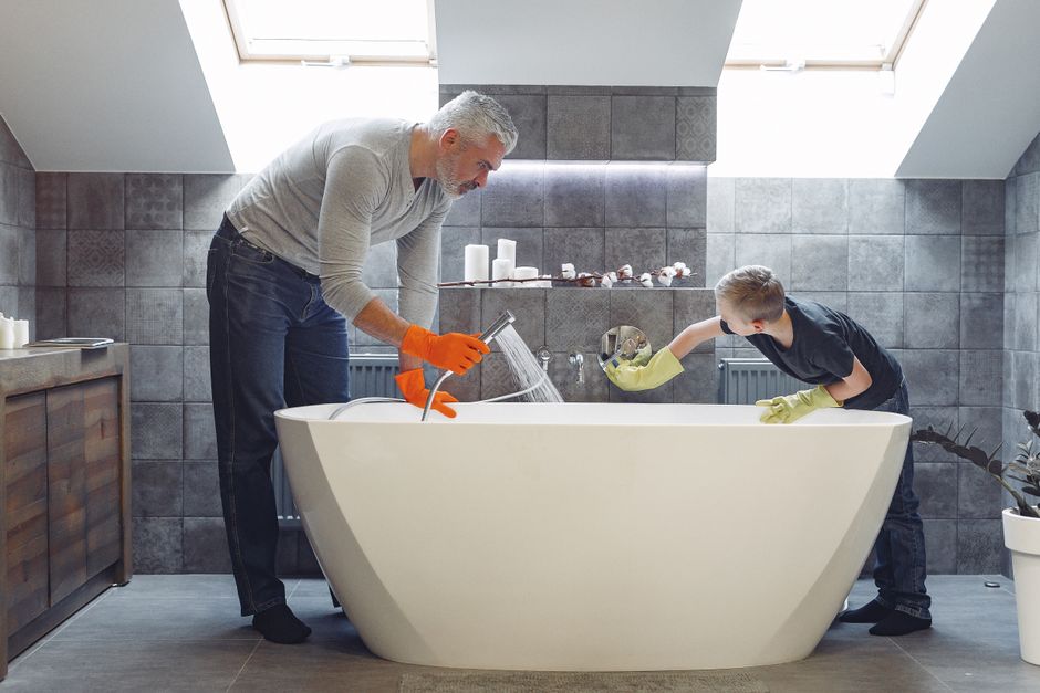Homem e criança limpando uma banheira em um banheiro com iluminação natural e parede de azulejos cinza, demonstrando uma atividade de cuidados domésticos.