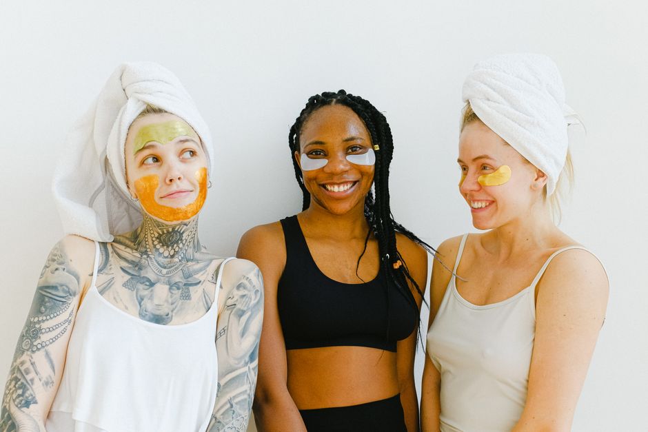 Três mulheres sorrindo com máscaras faciais e toalhas na cabeça em um ambiente de spa, demonstrando cuidados com a pele e bem-estar.