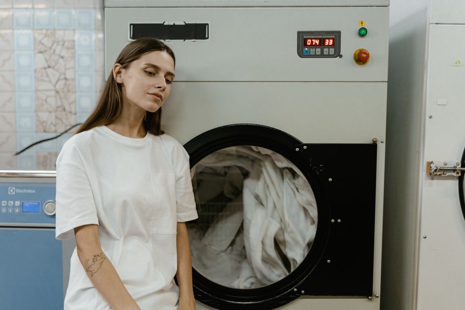 Mulher em ambiente industrial, próxima a uma máquina de lavar industrial, representando processos de lavanderia ou fabricação de roupas.