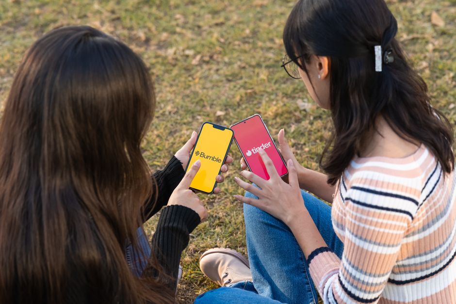 Duas jovens interagindo com seus smartphones ao ar livre, explorando aplicativos de namoro e redes sociais.