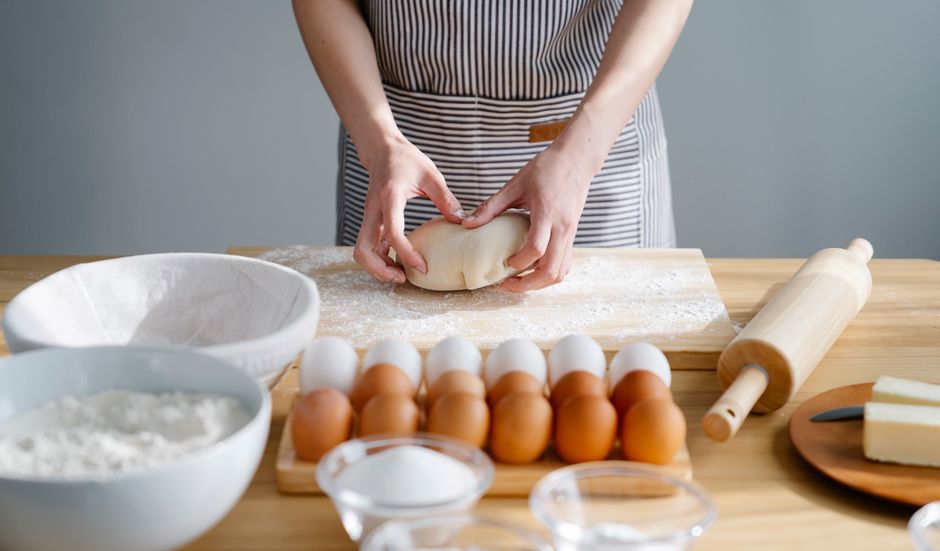 Imagem de uma pessoa preparando massa de pão ou massa caseira na cozinha, com ingredientes e utensílios ao redor, destacando a fabricação artesanal de alimentos.