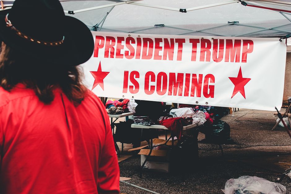 Pessoa observando uma faixa de campanha política apoiando o ex-presidente Trump em um evento ao ar livre.