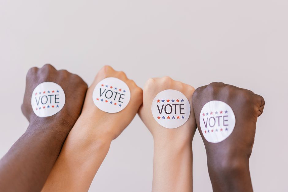 Imagem de mãos de diferentes cores com adesivos de votação, simbolizando diversidade e participação cívica.