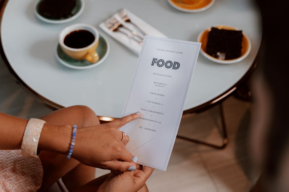 Pessoa segurando cardápio de restaurante em ambiente de café ou lanchonete, com diversas opções de comida e bebidas na mesa.