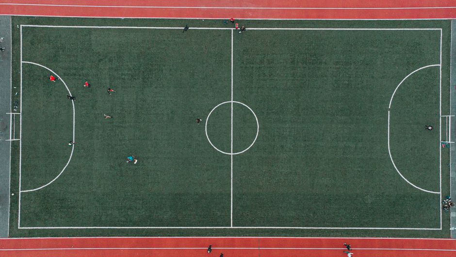 Imagem aérea de um campo de futebol com jogadores e a pista de corrida ao redor, capturando a dinâmica do esporte ao ar livre.
