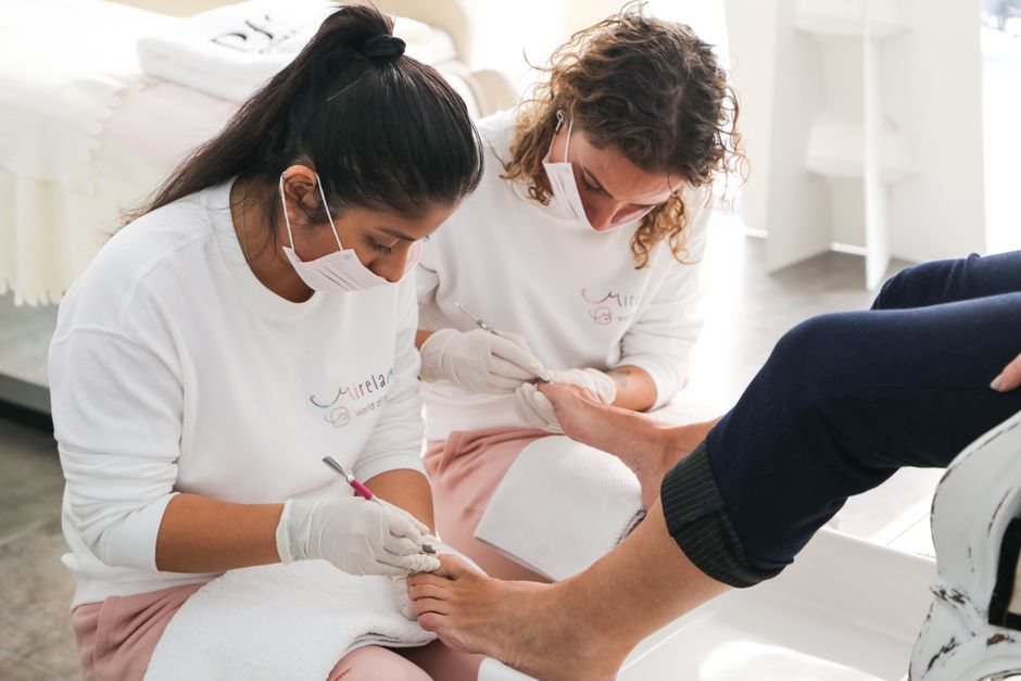 Duas profissionais realizando um procedimento de cuidados nos pés de um cliente em um ambiente clínico de estética ou podologia.