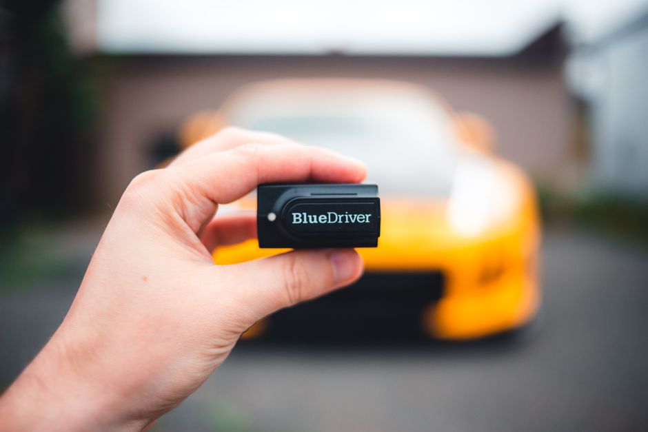 Imagem que mostra uma pessoa segurando um dispositivo de plug-in BlueDriver frente a um carro esportivo amarelo, relacionada à manutenção automotiva e diagnósticos de veículos.
