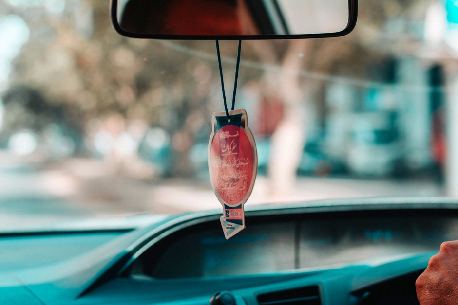 Imagem de um interior de carro visto do para-brisa, com um aviso de trânsito pendurado no espelho retrovisor e uma vista urbana desfocada ao fundo.