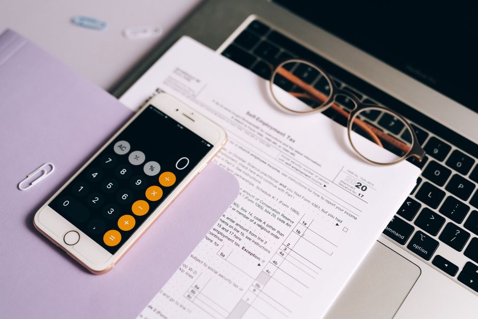 Imagem de uma área de trabalho com documentos fiscais, um laptop, um smartphone com calculadora e óculos, sugerindo planejamento financeiro ou análise tributária para conteúdo de blog sobre finanças pessoais ou empresariais.