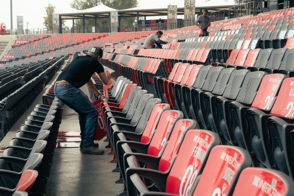 Pessoas limpando e arrumando cadeiras em um estádio, preparando o espaço para um evento esportivo ou cultural.