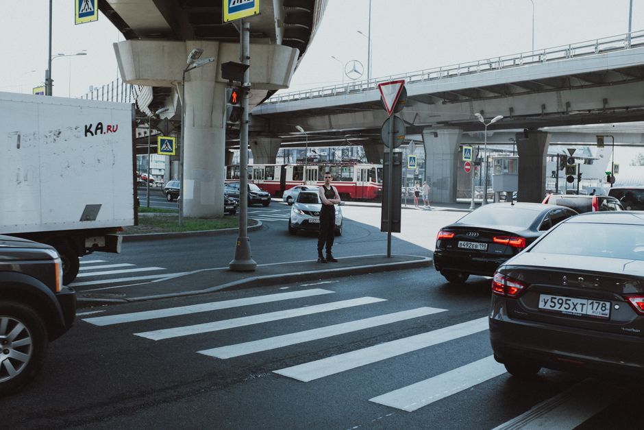 Imagem de um pedestre aguardando o momento de atravessar na faixa de segurança em uma movimentada rua urbana sob uma ponte elevada, com trânsito de carros e transporte público ao fundo.