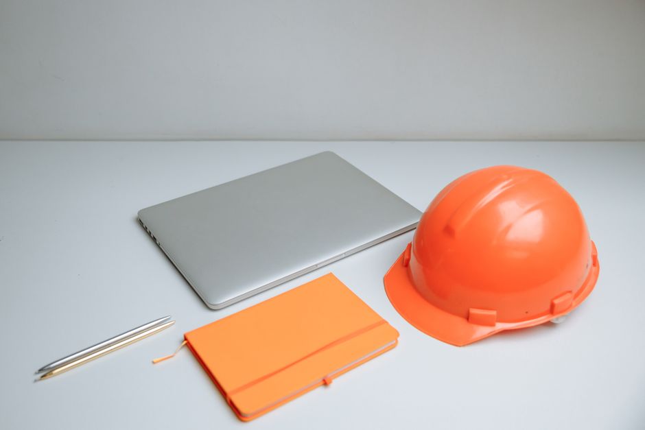 Imagem de um ambiente de trabalho que representa segurança e gestão de obras ou projetos, com objetos como capacete de segurança, notebook, caneta e agenda em uma mesa neutra.