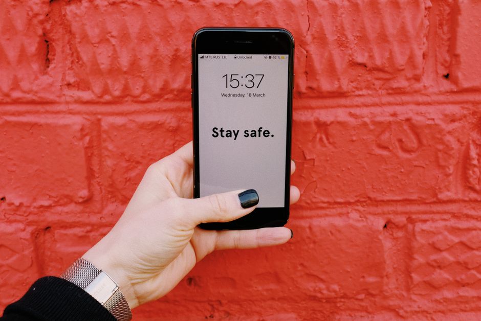 Pessoa segurando um smartphone com a mensagem 'Stay safe' em uma parede de tijolos vermelhos ao fundo, transmitindo uma mensagem de cuidado e segurança.